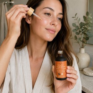 May include: A woman applying face serum from a brown glass bottle with a dropper. The bottle has an orange label with the words "FACE SERUM" and "MORNING + NIGHT". The serum is clear and the dropper is made of bamboo.