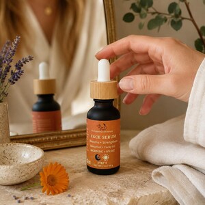 May include: A black glass face serum bottle with a bamboo cap and a dropper. The label is orange with white text that reads "FACE SERUM" and "Restore + Strengthen". The bottle is held near a mirror and a flower.