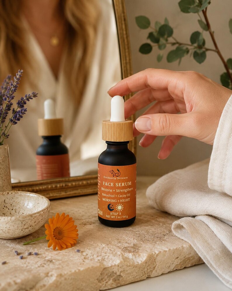 May include: A black glass face serum bottle with a bamboo cap and a dropper. The label is orange with white text that reads "FACE SERUM" and "Restore + Strengthen". The bottle is held near a mirror and a flower.