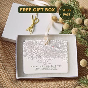May include: A rectangular white ceramic ornament featuring black topographic map lines and a small red heart. The text reads WHERE WE SAID SAID YES, 39.1178 N, 106.4453 W 09.2026, and BETTY & SCOTT - ENGAGED 09.22.2025. It hangs by a cream ribbon in a box.