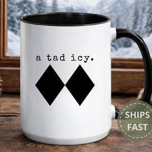 May include: White ceramic mug with a black interior and handle. The mug features the phrase "a tad icy" above two black diamond shapes. The mug is set against a wintery background.