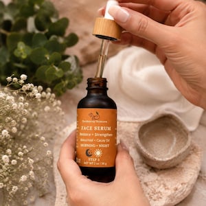 May include: A dark amber glass bottle of Outdoorsy Skincare Face Serum with a dropper. The label is orange with white text, including "Restore + Strengthen" and "Bakuchiol + Cacay Oil." The bottle is held near a small, light-colored bowl.