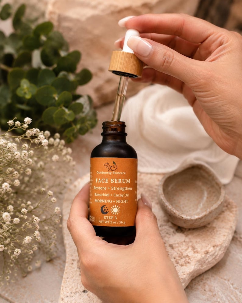May include: A dark amber glass bottle of Outdoorsy Skincare Face Serum with a dropper. The label is orange with white text, including "Restore + Strengthen" and "Bakuchiol + Cacay Oil." The bottle is held near a small, light-colored bowl.