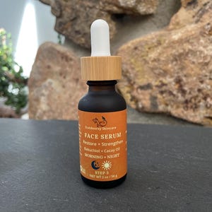 May include: A small, dark brown glass bottle of Outdoorsy Skincare Face Serum with a wooden cap and white dropper. The orange label includes text about restoring and strengthening the skin with Bakuchiol and Cacay Oil. The bottle is 2 oz / 56 g.