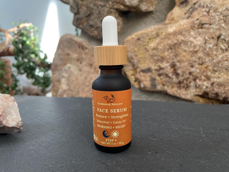 May include: A small, dark brown glass bottle of Outdoorsy Skincare Face Serum with a wooden cap and white dropper. The orange label includes text about restoring and strengthening the skin with Bakuchiol and Cacay Oil. The bottle is 2 oz / 56 g.