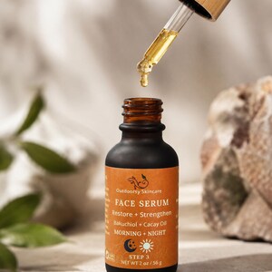 May include: A dark brown glass bottle of Outdoorsy Skincare Face Serum with a dropper dispensing the product. The label is orange with white text, including "Restore + Strengthen" and "Bakuchiol + Cacay Oil." The bottle contains 2 oz / 56 g of product.