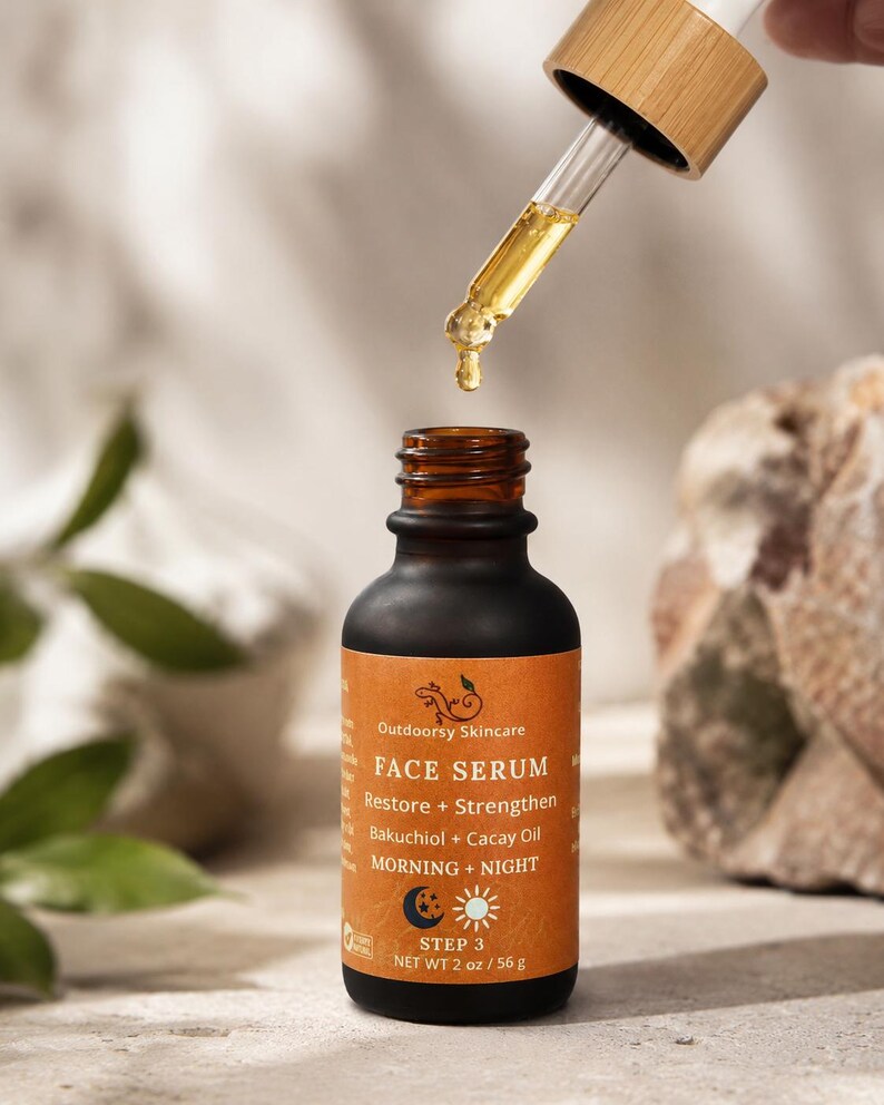 May include: A dark brown glass bottle of Outdoorsy Skincare Face Serum with a dropper dispensing the product. The label is orange with white text, including "Restore + Strengthen" and "Bakuchiol + Cacay Oil." The bottle contains 2 oz / 56 g of product.