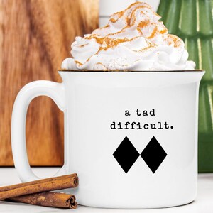 May include: White ceramic mug with the text "a tad difficult" and two black diamond shapes. The mug is filled with a whipped cream topping dusted with cinnamon. Two cinnamon sticks rest in front of the mug.