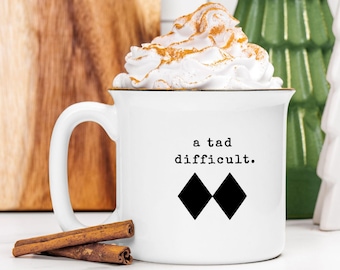 May include: White ceramic mug with the text "a tad difficult" and two black diamond shapes. The mug is filled with a whipped cream topping dusted with cinnamon. Two cinnamon sticks rest in front of the mug.