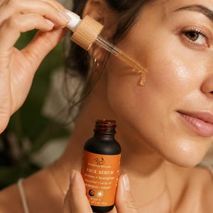 May include: A woman applying face serum from a small brown bottle with an orange label. The label reads "Outdoorsy Skincare FACE SERUM Restore + Strengthen Bakuchiol + Cacay Oil MORNING + NIGHT STEP 3 WT 2 oz / 56 g". The serum is dispensed with a dropper.