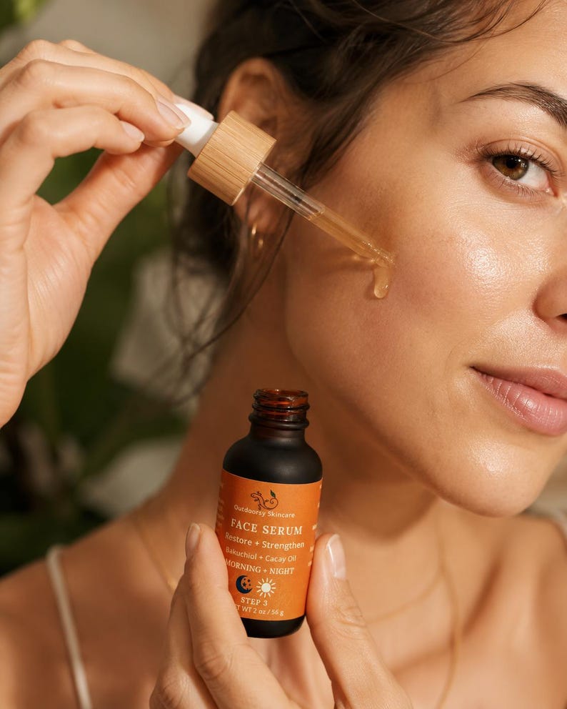 May include: A woman applying face serum from a small brown bottle with an orange label. The label reads "Outdoorsy Skincare FACE SERUM Restore + Strengthen Bakuchiol + Cacay Oil MORNING + NIGHT STEP 3 WT 2 oz / 56 g". The serum is dispensed with a dropper.