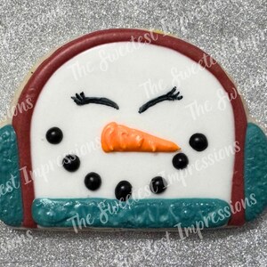 May include: A snowman-shaped cookie with white icing, black dot eyes, and an orange carrot nose. The cookie has a red border and teal earmuffs. The text "The Sweetest Impressions" is visible on the cookie.