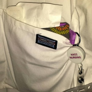 May include: A white scrub top with a pocket that has a Cherokee Workwear label and a white badge with the text "HACC NURSING" on it.