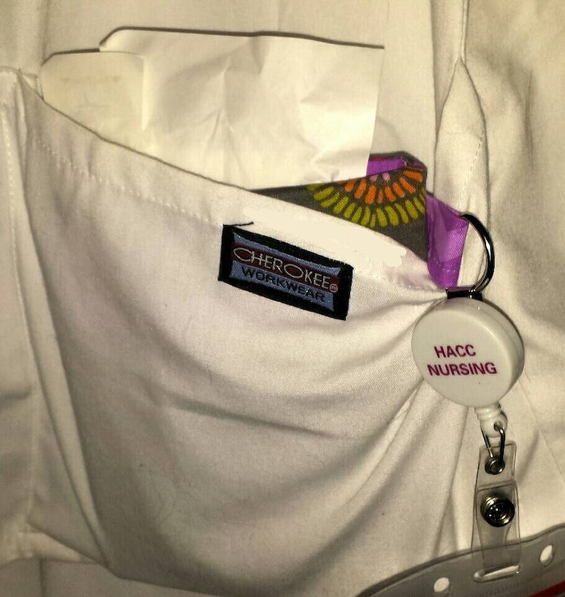 May include: A white scrub top with a pocket that has a Cherokee Workwear label and a white badge with the text "HACC NURSING" on it.