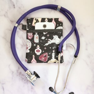 May include: A purple stethoscope with a black and white fabric pouch with a potion and magic themed print. A white smartphone is in the pouch.