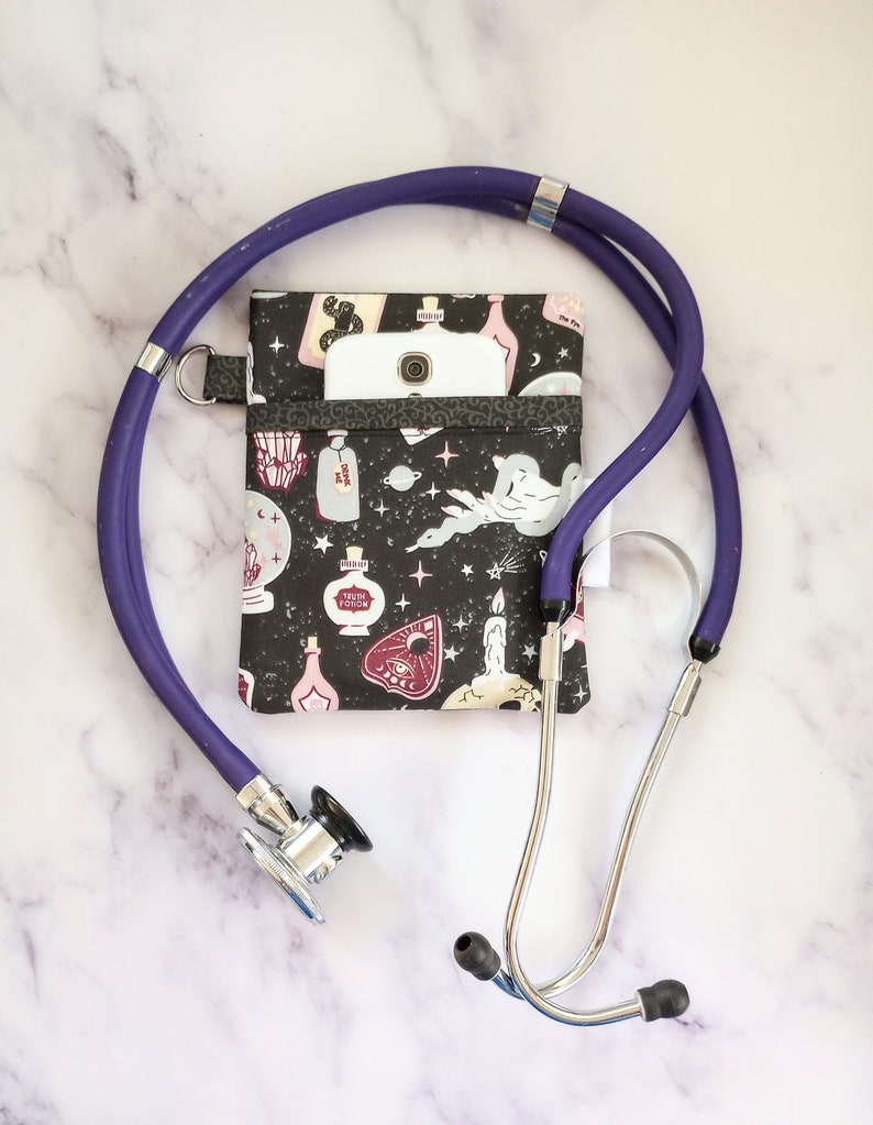 May include: A purple stethoscope with a black and white fabric pouch with a potion and magic themed print. A white smartphone is in the pouch.