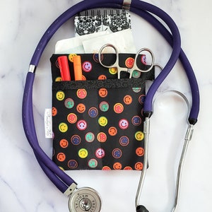 May include: A black fabric pouch with a smiley face pattern, containing a small notebook, a pair of scissors, and two pens. A purple stethoscope is wrapped around the pouch. The stethoscope has a silver head with the word "Prestige Medical" printed on it.