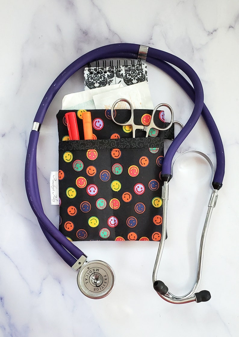 May include: A black fabric pouch with a smiley face pattern, containing a small notebook, a pair of scissors, and two pens. A purple stethoscope is wrapped around the pouch. The stethoscope has a silver head with the word "Prestige Medical" printed on it.