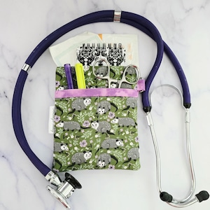 May include: A green fabric pouch with a purple trim and a pattern of opossums. The pouch is attached to a stethoscope with a dark blue tube and silver earpieces.