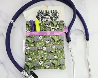 May include: A green fabric pouch with a purple trim and a pattern of opossums. The pouch is attached to a stethoscope with a dark blue tube and silver earpieces.