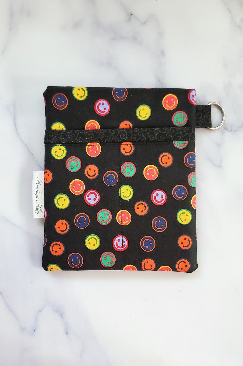 May include: Black fabric pouch with a colorful smiley face pattern. The pouch has a zipper closure and a metal ring attached to the top. The pouch is labeled "Amethyst Alley".