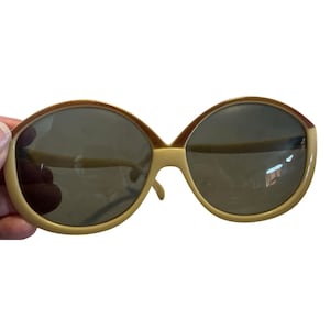 May include: Oversized sunglasses with a light brown frame and dark lenses. The sunglasses have a rounded shape and a thick frame. The lenses are a dark gray color. The sunglasses are held by a hand.