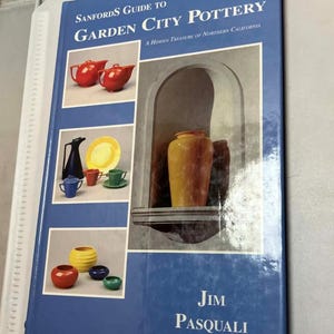 May include: A blue book titled "Sanfords Guide to Garden City Pottery" with images of pottery pieces. The cover features red teapots, a black pitcher, yellow plates, and colorful bowls. The author's name, Jim Pasquali, is printed at the bottom.