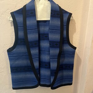 May include: A sleeveless vest featuring a blue gradient pattern with horizontal stripes and dark geometric diamond motifs. The garment has a wide shawl collar and is finished with solid black trim along the front opening and armholes.