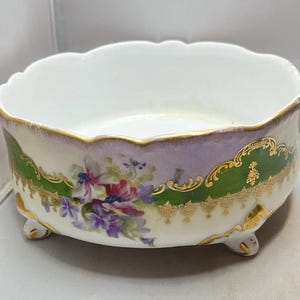 May include: A white porcelain bowl with a scalloped rim, featuring a floral design in purple, pink, and green. The bowl has a gold trim and rests on three small feet. The exterior has a green and purple band.