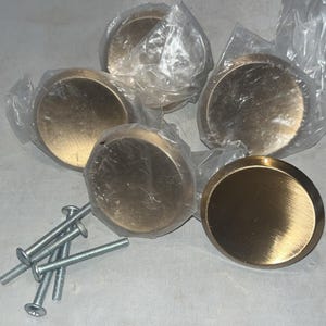 May include: A set of five round, antique brass cabinet knobs, each in a clear plastic wrapper. The knobs have a brushed finish and are accompanied by a set of silver screws for installation. These hardware pieces are ideal for updating furniture.