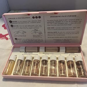 May include: A pink rectangular case containing twelve small glass bottles of foundation and powder, with instructional text printed on the inside lid. The set includes labels for fair and deep finishes, along with a small white applicator tool.