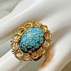 May include: A vintage gold-toned ring featuring an oval-shaped, speckled turquoise stone. The stone is set within a decorative filigree frame. The ring's design is ornate and classic, suitable for a jewelry collection.