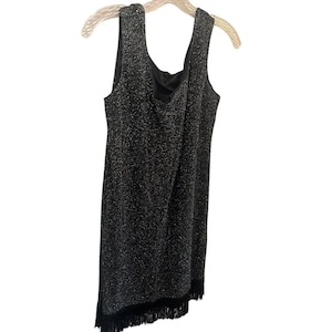 May include: A sleeveless black dress featuring a shimmering silver metallic thread throughout the fabric. The garment has a scoop neckline and a straight silhouette, finished with a row of black fringe trim along the bottom hemline.