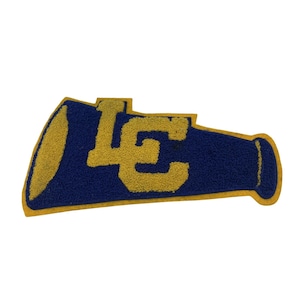May include: A blue and gold chenille patch shaped like a megaphone featuring the letters LC in a bold, block font. The patch has a textured, fuzzy surface with a gold border outlining the megaphone shape and the central letters.