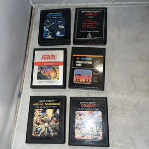 May include: Six rectangular black Atari 2600 video game cartridges arranged in two columns. The labels feature titles including Asteroids, Combat, Centipede, Commando Raid, and Missile Command, with colorful graphic illustrations and white text.