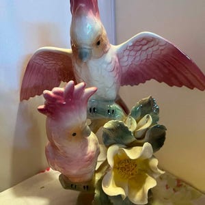 May include: A porcelain figurine featuring two pink and white cockatoos perched on a branch. The top bird has its wings spread wide, while the lower bird sits near a cluster of large, cream-colored flowers with yellow centers and green leaves.