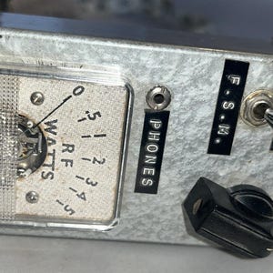 May include: A vintage silver-colored radio frequency (RF) power meter with a clear display and a needle indicating watts. The device has a switch labeled "WATTS" and a knob. Additional labels include "PHONES" and "F.S.M."