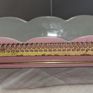 May include: A rectangular ceramic serving dish with a scalloped rim and a light pink exterior. The dish is held within a gold-toned metal wire frame stand that features a decorative filigree border, two side handles, and four thin legs.