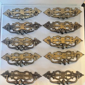 May include: A collection of ten antique brass drawer pulls. Each pull features an ornate, symmetrical design with a central handle and decorative detailing. The pulls have a vintage aesthetic, with a mix of gold and darker tones.