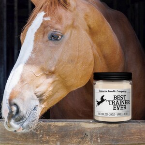 May include: A brown and white horse looks out of a wooden stall door. A black candle jar with a white label reads "Best Trainer Ever" in front of the horse.