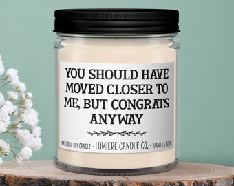 May include: A white soy candle in a clear glass jar with a black lid. The candle has a label that reads "You should have moved closer to me, but congrats anyway".