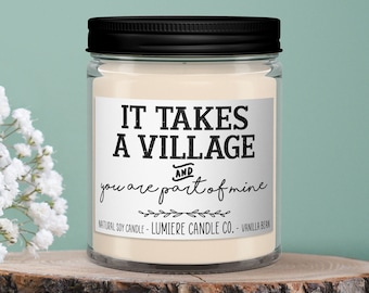 May include: A glass jar candle with a black lid and a white label that reads "It takes a village and you are part of mine". The candle is sitting on a wooden slice and surrounded by white baby's breath flowers.
