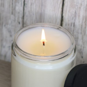 Puede incluir: Una vela en un tarro de vidrio transparente con cera blanca y una mecha encendida. La vela est&aacute; sobre una superficie de madera.