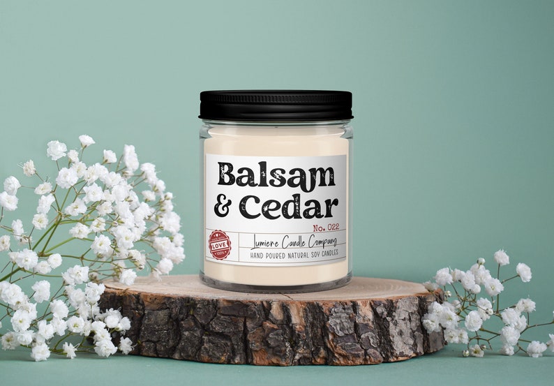 Puede incluir: Una vela de vidrio en un frasco con tapa negra y una etiqueta blanca. La etiqueta dice "Balsam & Cedar" y "Lumiere Candle Company" con un sello rojo de "Love".