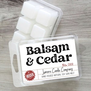 Puede incluir: Un recipiente de pl&aacute;stico transparente con seis pastillas de cera blanca. El recipiente tiene una etiqueta que dice "Balsam & Cedar" y "Lumiere Candle Company". La etiqueta tambi&eacute;n incluye el texto "No. 022" y "Hand Poured Natural Soy Wax Melt".