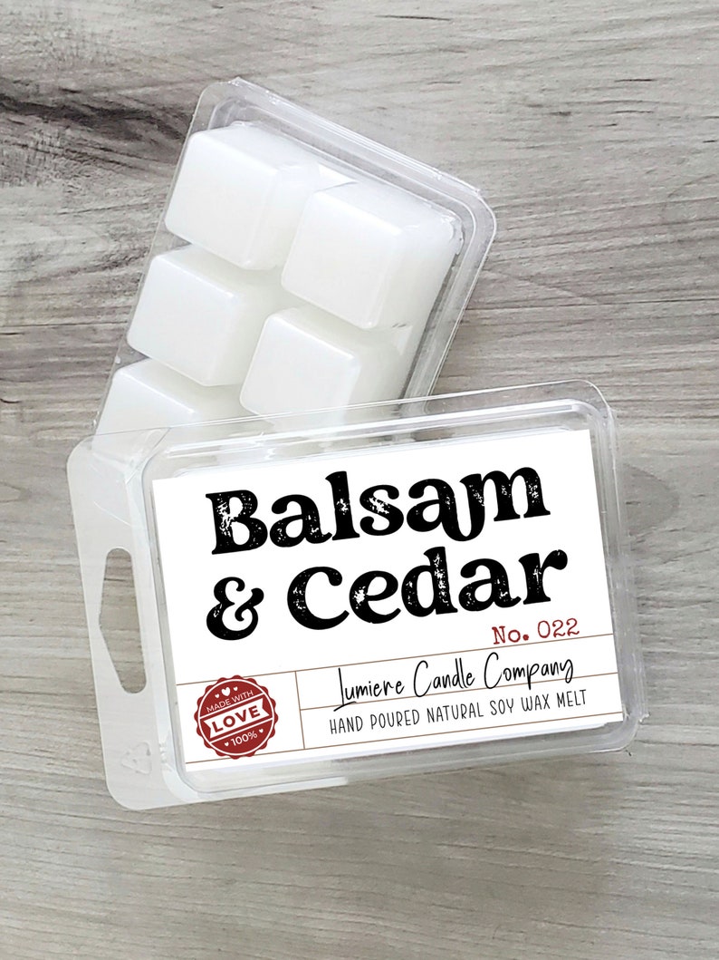 Puede incluir: Un recipiente de pl&aacute;stico transparente con seis pastillas de cera blanca. El recipiente tiene una etiqueta que dice "Balsam & Cedar" y "Lumiere Candle Company". La etiqueta tambi&eacute;n incluye el texto "No. 022" y "Hand Poured Natural Soy Wax Melt".