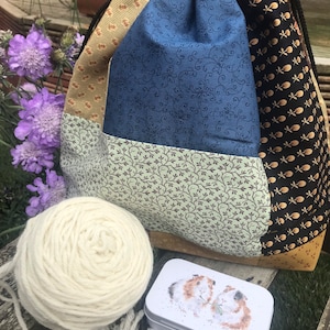 Peut inclure: Un sac à cordon en patchwork avec un motif floral bleu, vert et or. Le sac est rempli d'une pelote de laine blanche et d'une petite boîte en étain avec une image de deux cochons d'Inde.