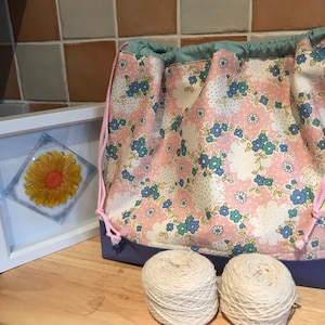 May include: A pink and white floral drawstring bag with a blue lining. Two balls of white yarn are in front of the bag.