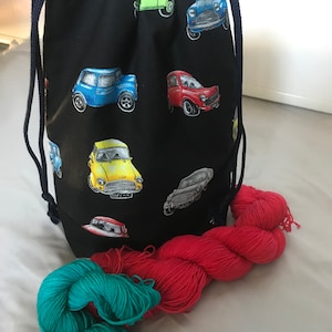 Può includere: Una borsa a cordoncino nera con un motivo di mini auto colorate. La borsa è piena di due gomitoli di filo, uno rosso e uno turchese.