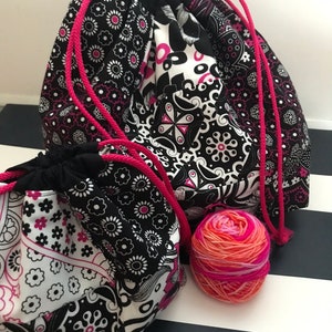 May include: Two black and white patterned drawstring bags with pink drawstrings. The larger bag is partially open and a ball of pink, orange and white yarn is sitting in front of it.